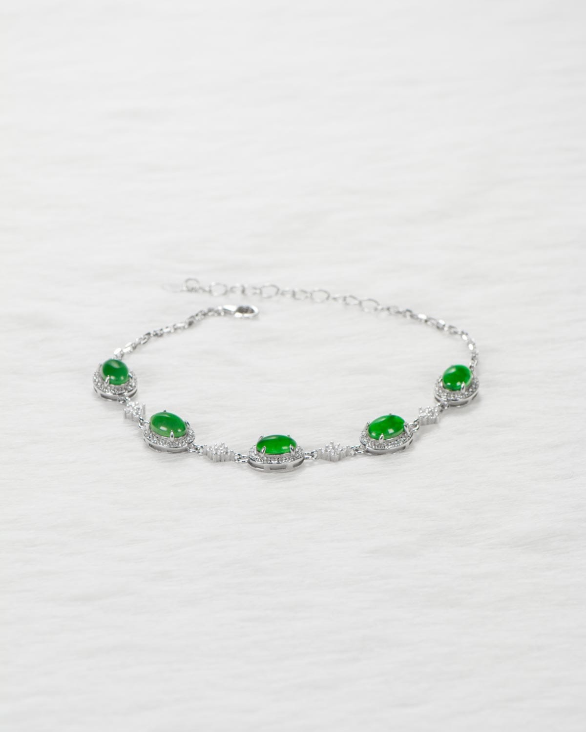 A silver bracelet with five oval green gemstones, each surrounded by small clear stones, displayed on a white textured surface.