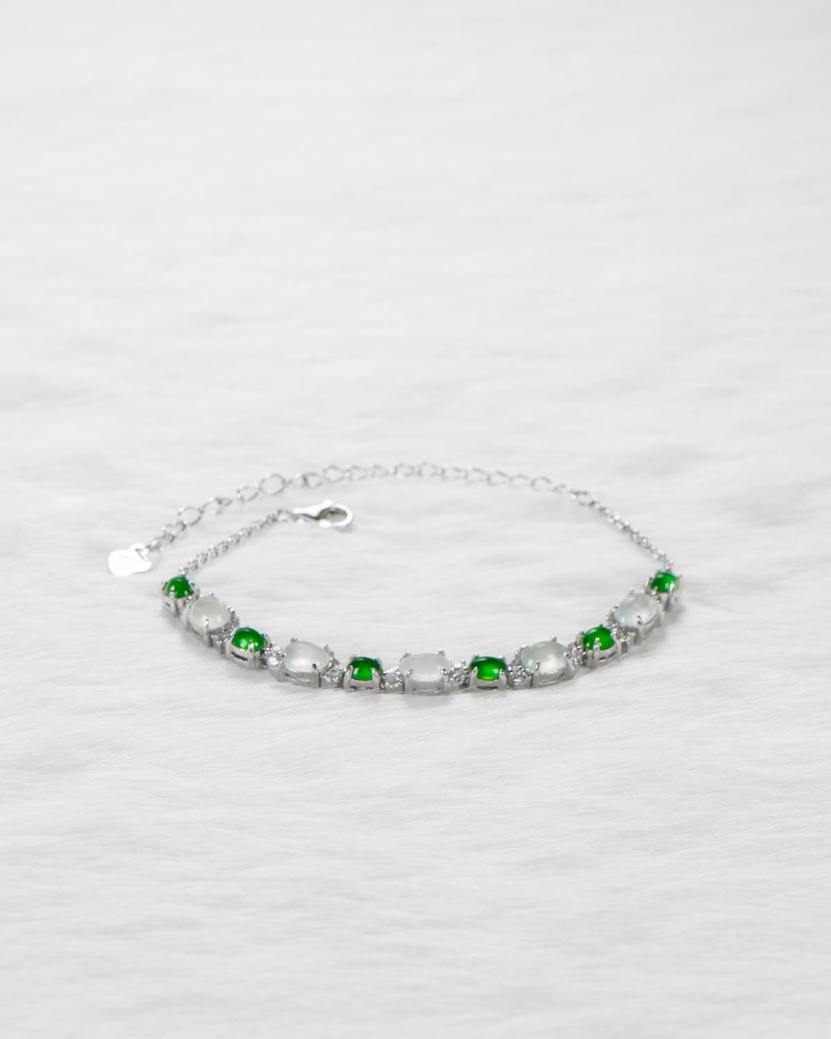 A delicate silver bracelet with alternating green and clear gemstones is displayed on a soft, white textured surface.