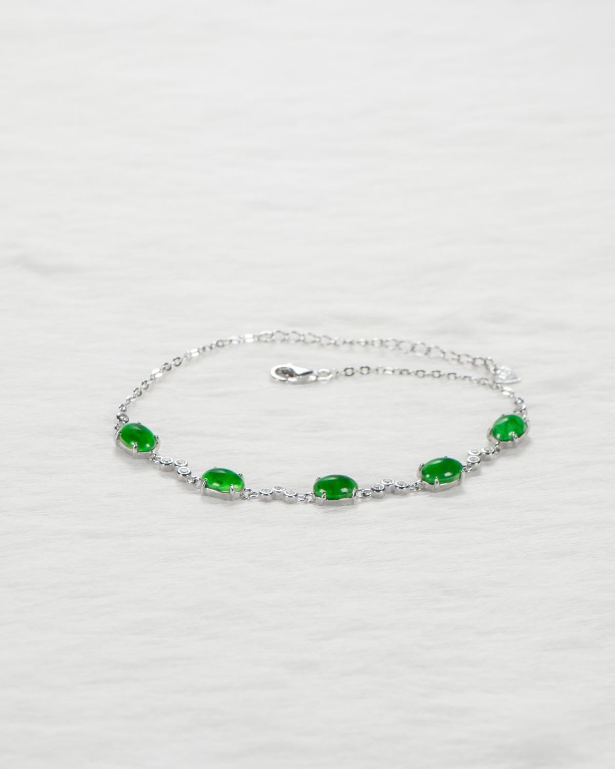 A delicate silver bracelet with five oval green gemstones evenly spaced along the chain, displayed on a light, textured surface.