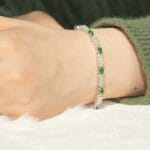 A close-up of a persons wrist wearing a delicate bracelet with alternating green and clear beads. The person is dressed in a green, fuzzy long-sleeve top.