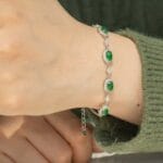 A hand wearing a silver bracelet with oval green gemstones and small clear stones, shown against a green knitted sleeve background.