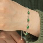 A close-up of a hand wearing a silver bracelet adorned with green gemstones and small clear crystals, against a knit green sweater background.