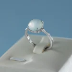 A silver ring with a round, milky white gemstone is displayed on a cream-colored ring holder against a light blue background.