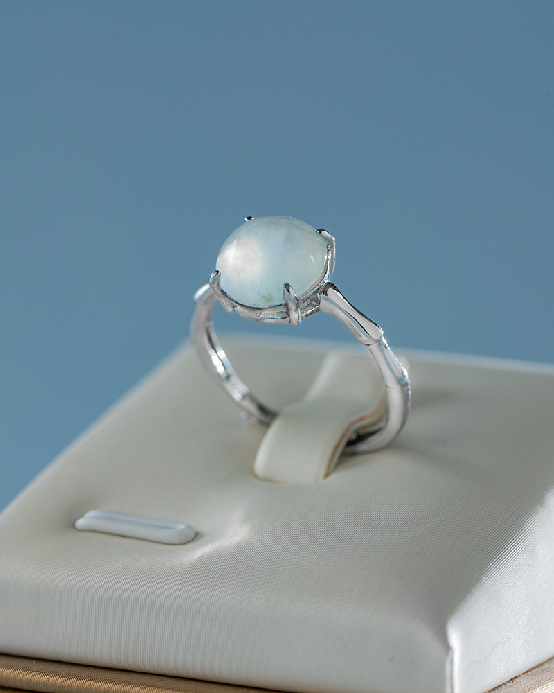 DSC_0182 拷贝 A silver ring with a round, milky white gemstone is displayed on a cream-colored ring holder against a light blue background.