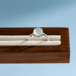 A silver ring with a round, translucent gemstone is displayed on a cream-colored, velvet-lined wooden jewelry stand against a light blue background.