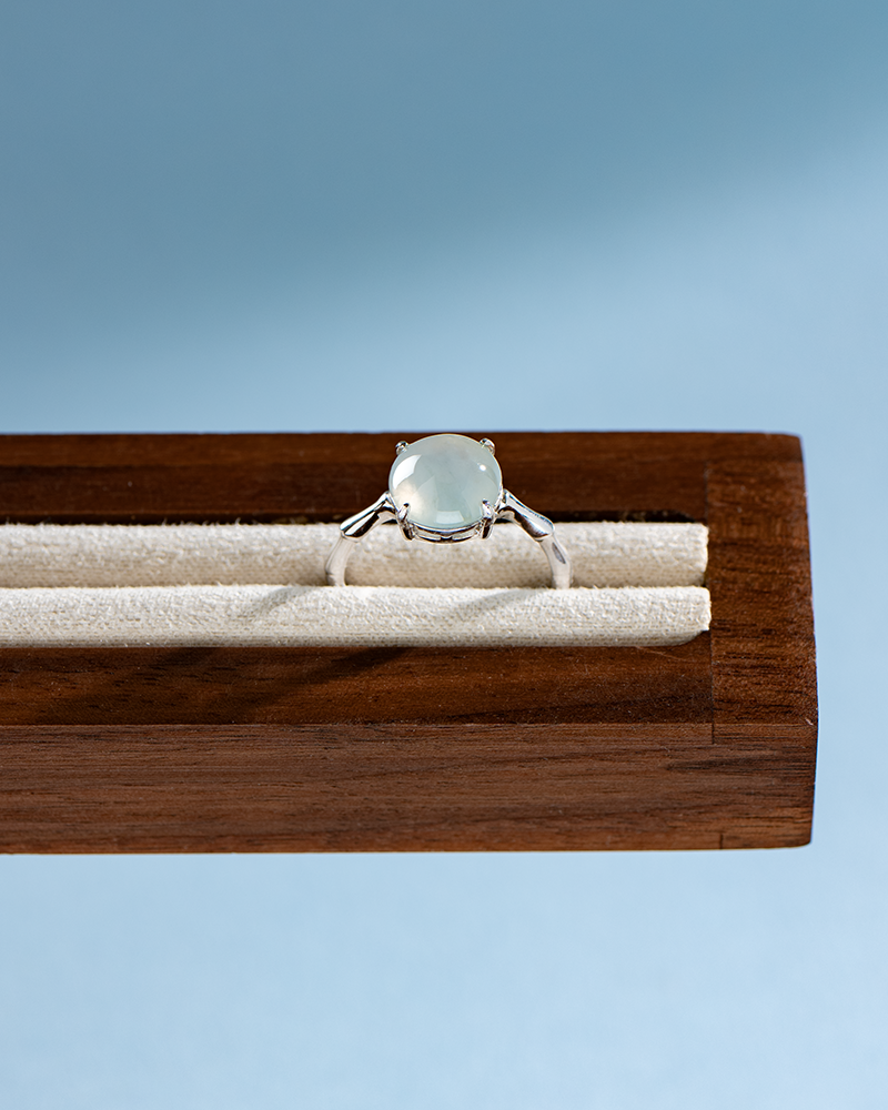 DSC_0184 拷贝 A silver ring with a round, translucent gemstone is displayed on a cream-colored, velvet-lined wooden jewelry stand against a light blue background.