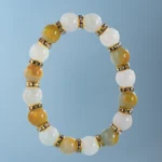 A beaded bracelet with alternating white, light green, and amber round stones, separated by gold spacers with embedded rhinestones, displayed on a light blue background.