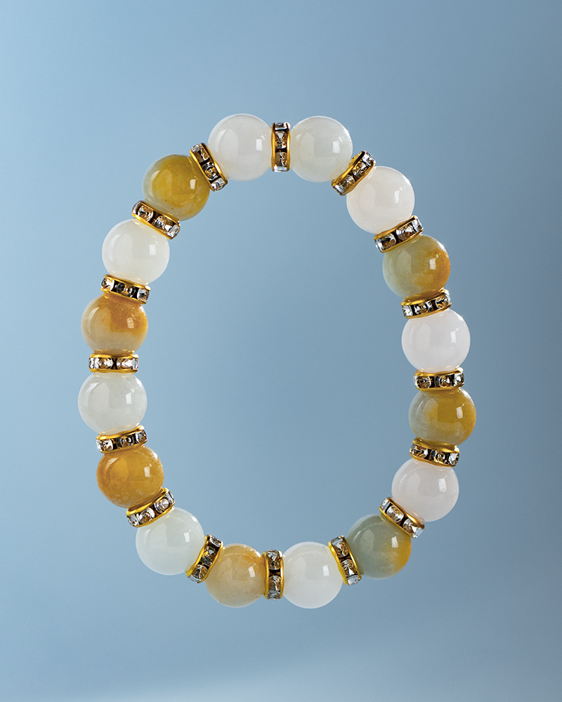 A beaded bracelet with alternating white, light green, and amber round stones, separated by gold spacers with embedded rhinestones, displayed on a light blue background.