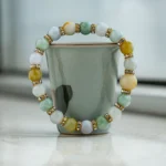 A beaded bracelet with multicolored stones and gold accents is balanced upright around a pale green ceramic cup on a reflective surface by a window.