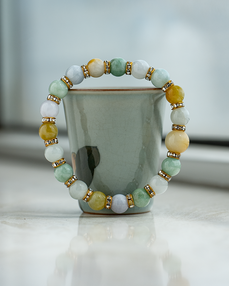 A beaded bracelet with multicolored stones and gold accents is balanced upright around a pale green ceramic cup on a reflective surface by a window.