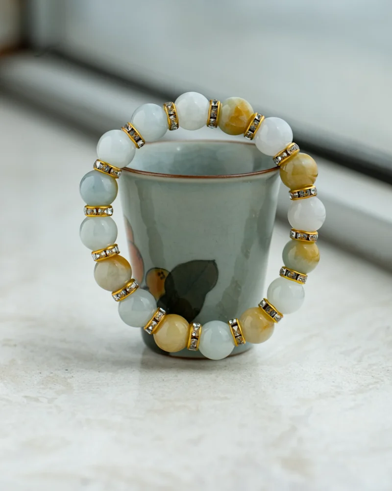 A Greenish White and Brownish Yellow Jadeite Bracelet featuring spherical beads with faceted accents is displayed draped over a ceramic cup with a floral design on a light surface.