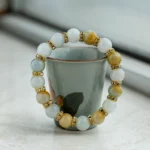 A beaded bracelet with alternating white and yellow beads, separated by gold spacers with rhinestones, is draped over a small ceramic cup with a floral design on a light surface.