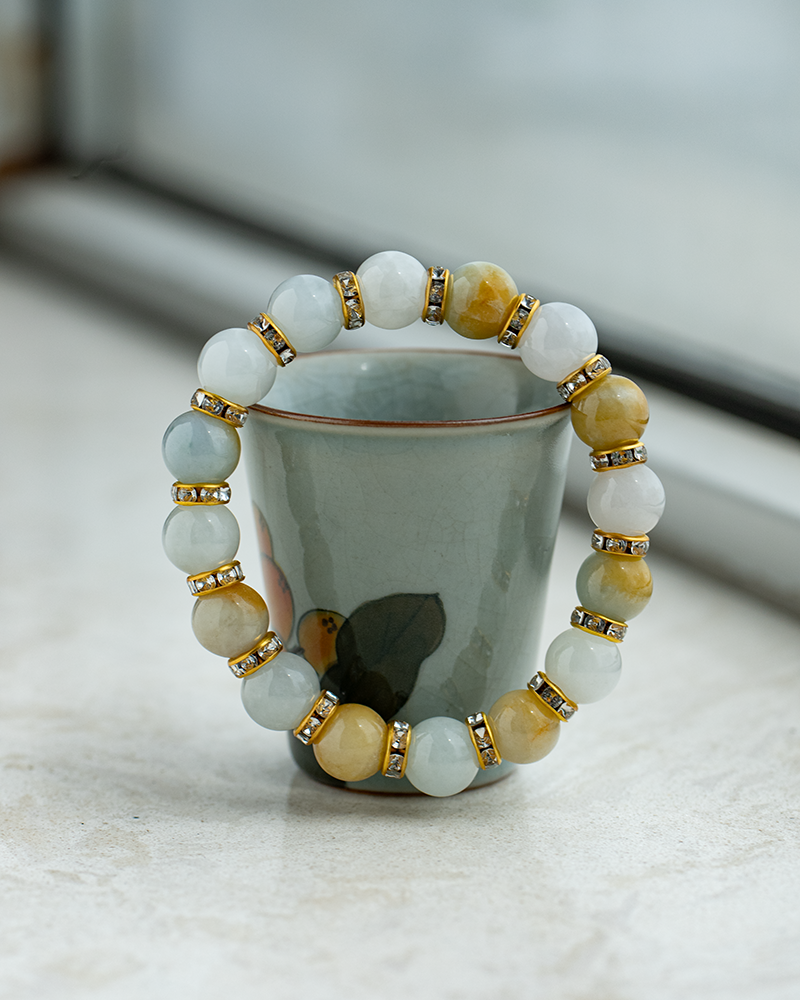 A beaded bracelet with alternating white and yellow beads, separated by gold spacers with rhinestones, is draped over a small ceramic cup with a floral design on a light surface.