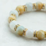 A close-up of a beaded bracelet with polished stones in shades of white, light blue, and yellow, separated by gold spacers with small, clear gemstones, on a light textured surface.