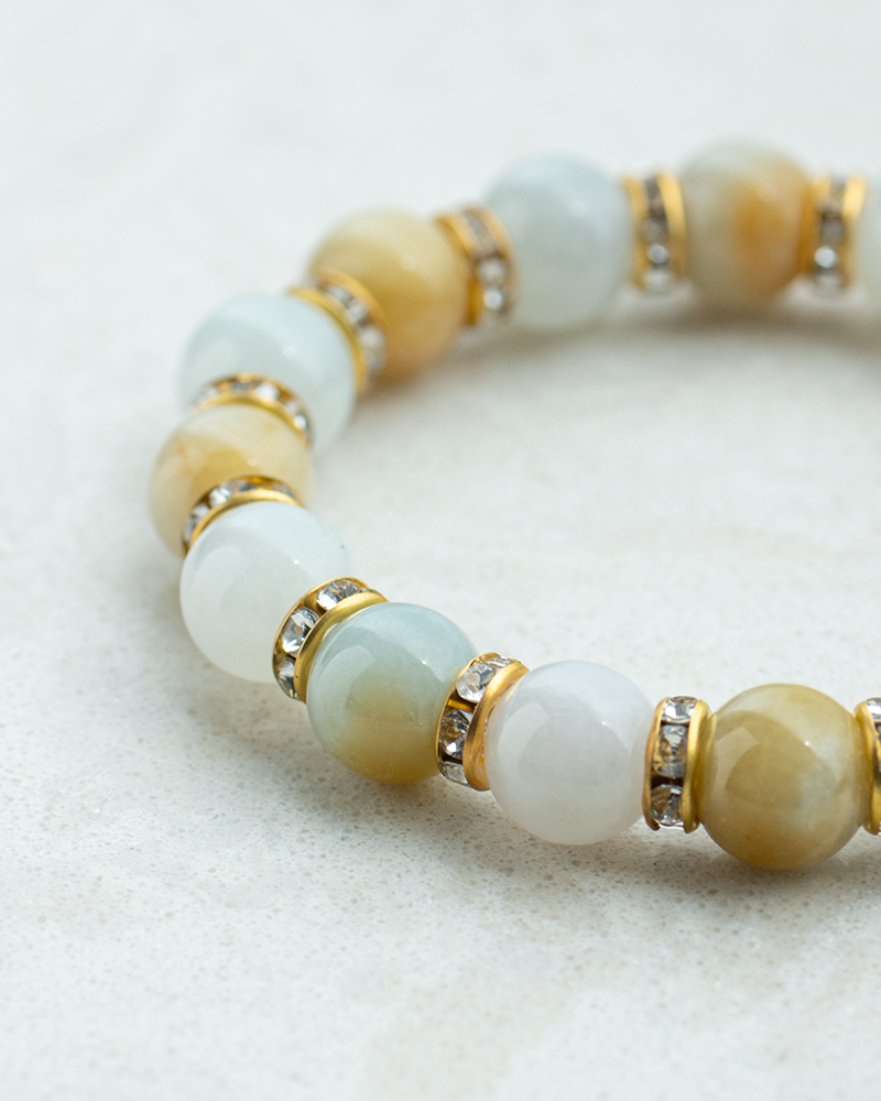 A close-up of a beaded bracelet with polished stones in shades of white, light blue, and yellow, separated by gold spacers with small, clear gemstones, on a light textured surface.