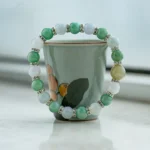 A beaded bracelet with green, white, and yellow stones separated by small metallic spacers is displayed around a small ceramic cup with a painted flower design, set on a light surface.