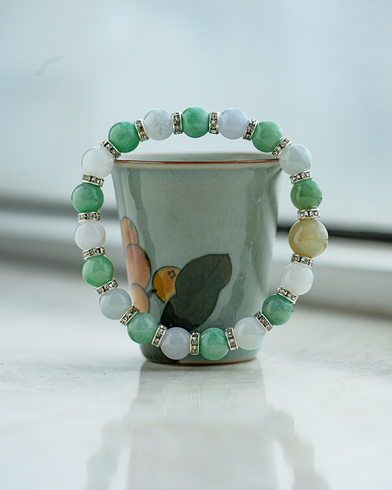 A beaded bracelet with green, white, and yellow stones separated by small metallic spacers is displayed around a small ceramic cup with a painted flower design, set on a light surface.