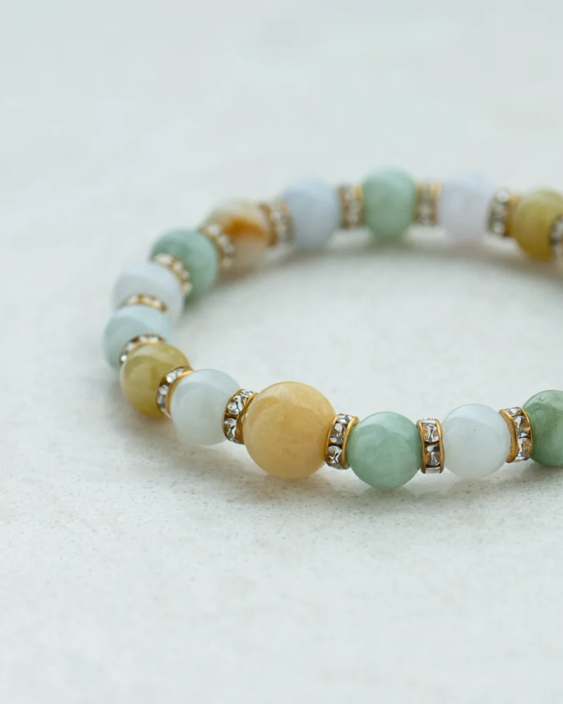 A close-up of the Light Green Jadeite Beaded Bracelet, showcasing spherical light green jadeite beads accented with round brownish yellow stones and separated by gold-tone spacers adorned with embedded crystals, all arranged on a light, textured surface.