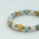 A beaded bracelet with round jade stones in shades of green, yellow, and white, separated by gold spacers with small crystals, displayed on a light, textured surface.