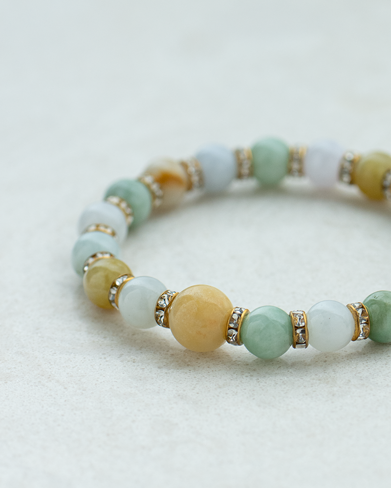 A beaded bracelet with round jade stones in shades of green, yellow, and white, separated by gold spacers with small crystals, displayed on a light, textured surface.