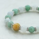 A beaded bracelet with green, white, and one prominent yellow bead, separated by silver spacers, displayed on a light textured surface.