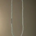 A long necklace made of small, light blue beads with a decorative silver clasp, displayed against a plain brown gradient background.