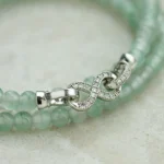 A close-up of a green beaded bracelet with a silver infinity-shaped clasp adorned with small crystals, displayed on a light beige surface.