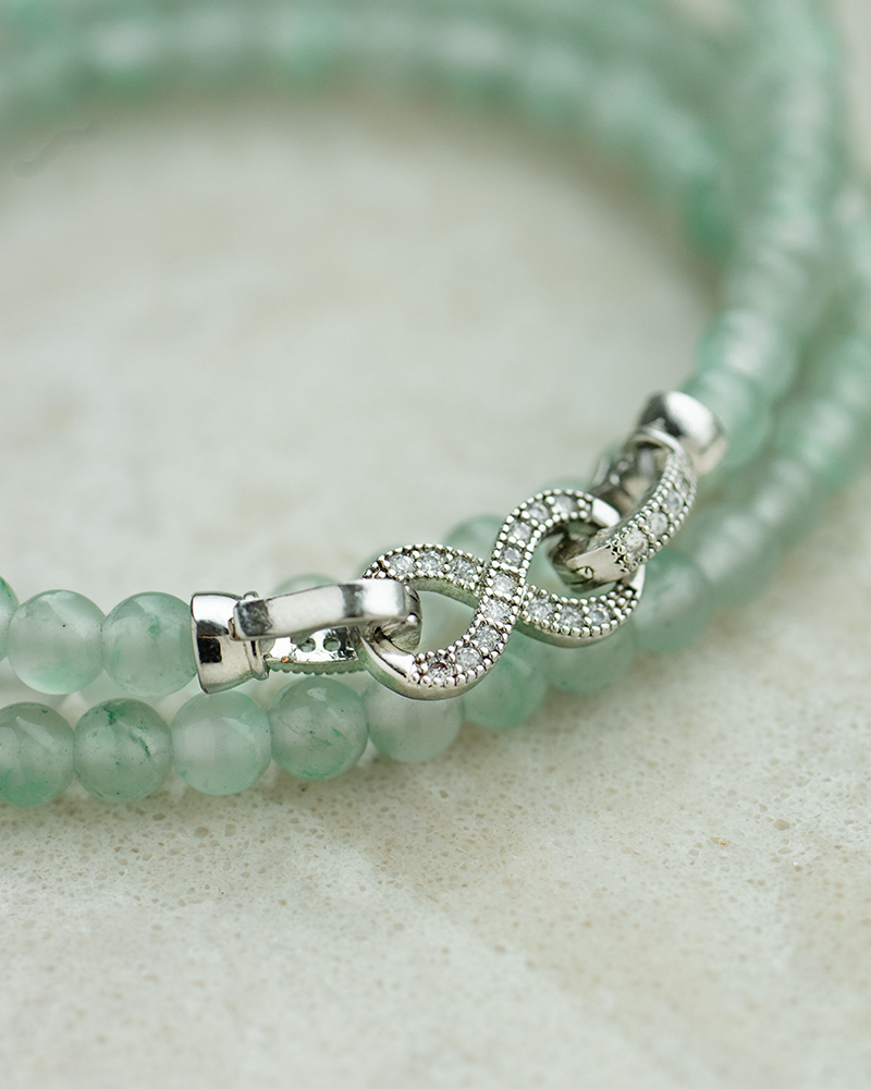 A close-up of a green beaded bracelet with a silver infinity-shaped clasp adorned with small crystals, displayed on a light beige surface.