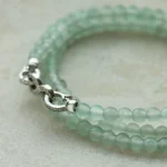 A close-up of a double-stranded bracelet made of translucent light green beads with a silver clasp, resting on a light textured surface.