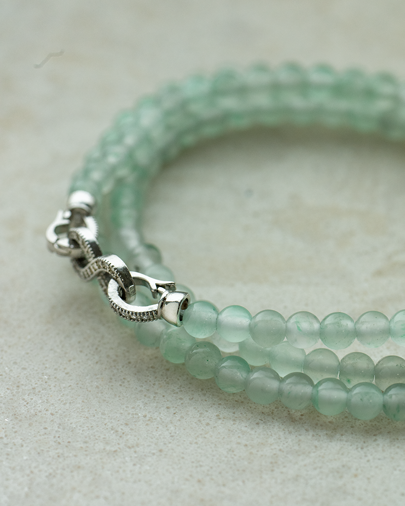 A close-up of a double-stranded bracelet made of translucent light green beads with a silver clasp, resting on a light textured surface.