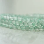 A close-up of three strands of pale green, translucent jade beads with subtle darker green patterns, resting on a soft, light-colored surface.