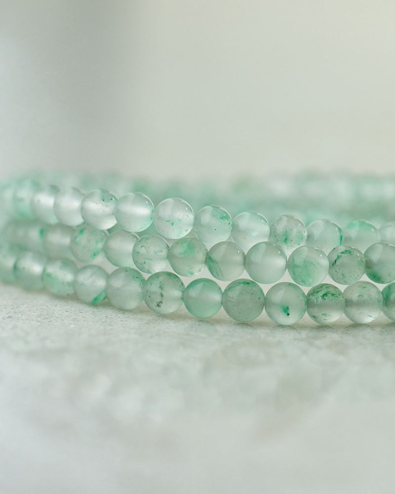 A close-up of three strands of pale green, translucent jade beads with subtle darker green patterns, resting on a soft, light-colored surface.