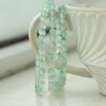 A string of translucent green beads is draped over the handle of a white, textured ceramic cup sitting on a light-colored surface.