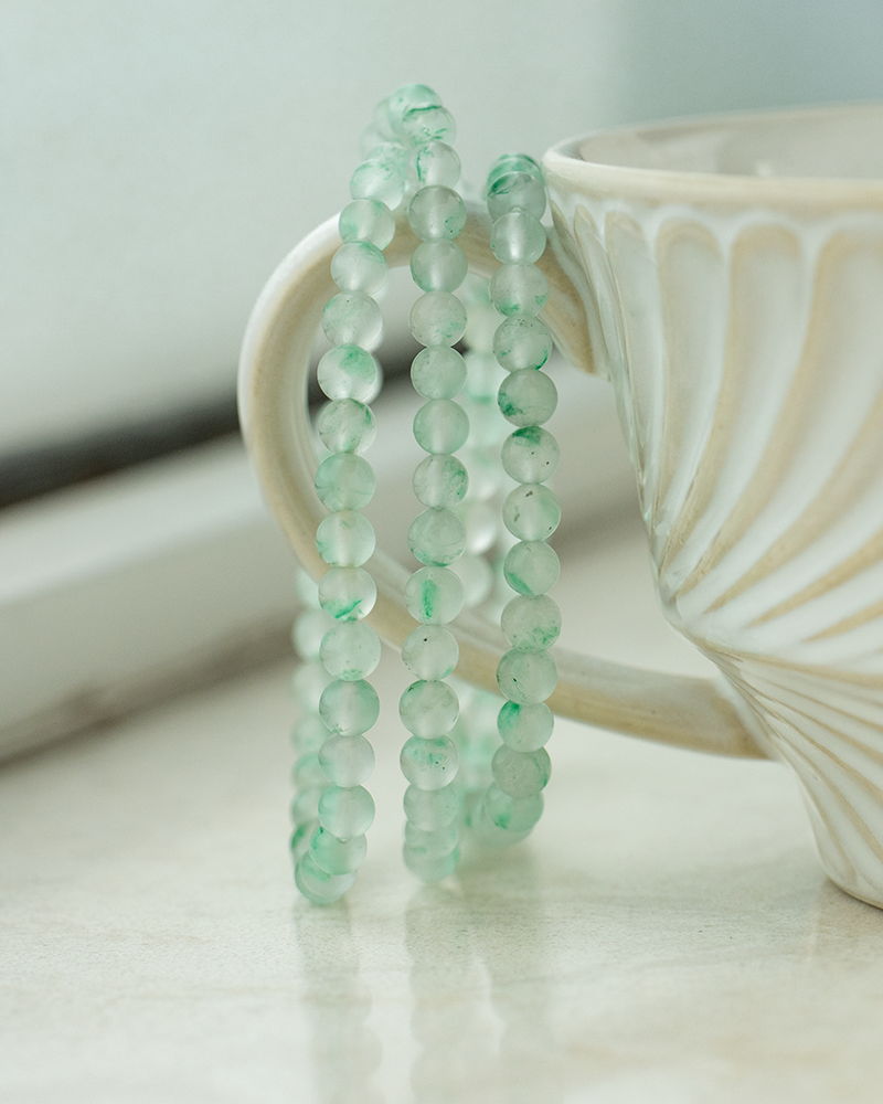 A string of translucent green beads is draped over the handle of a white, textured ceramic cup sitting on a light-colored surface.