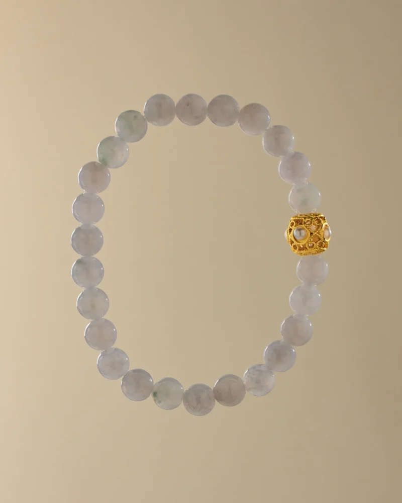 The Greenish-White Jadeite Bead Bracelet — Spherical Beads with Warm-Tone Accent, showcasing light gray jadeite beads accented by a single ornate gold bead, is elegantly displayed against a plain beige background.