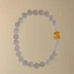 A beaded bracelet made of light gray stones with a single ornate gold bead, displayed on a plain beige background.