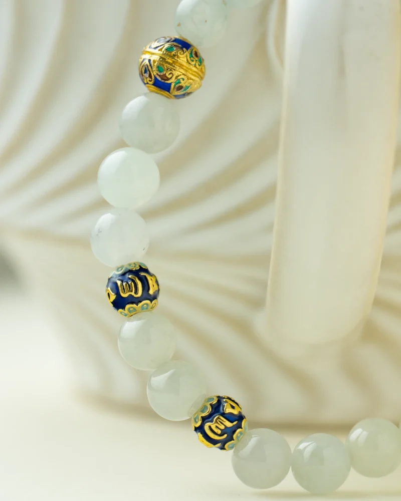 A close-up of the Light Green Jadeite Bead Bracelet — Spherical Beads with Warm Accents, featuring pale green jadeite beads and three ornate blue-and-gold enamel beads, draped over a white, textured ceramic mug.