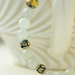 A close-up of the Light Green Jadeite Bead Bracelet—spherical beads with warm accents—displayed on the handle of a textured white ceramic cup.