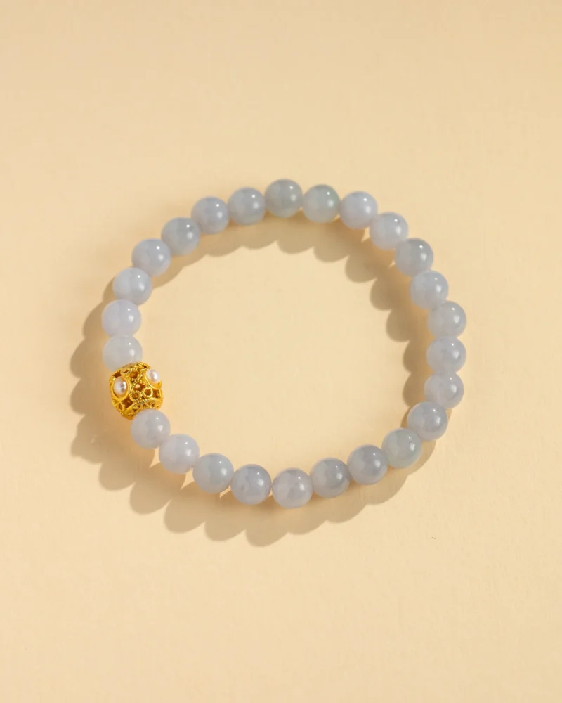 A Greenish-White Jadeite Bead Bracelet — Spherical Beads with Warm-Tone Accent, displayed in a circular arrangement on a light beige background. The pale blue-green hues of the jadeite beads are complemented by a single ornate gold accent adorned with tiny pearls, adding an elegant and refined touch to the delicate design.