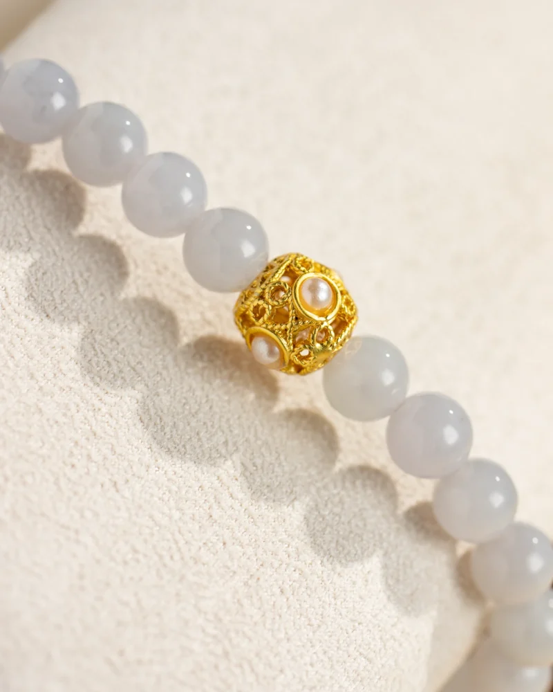 A close-up of the Greenish-White Jadeite Bead Bracelet — Spherical Beads with Warm-Tone Accent, showcasing pale gray round jadeite beads and a single ornate gold accent adorned with delicate pearls, resting on a textured beige surface.