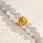 A close-up of a beaded bracelet with smooth, pale gray beads and a single ornate gold bead featuring small pearls, displayed on a textured light beige surface.