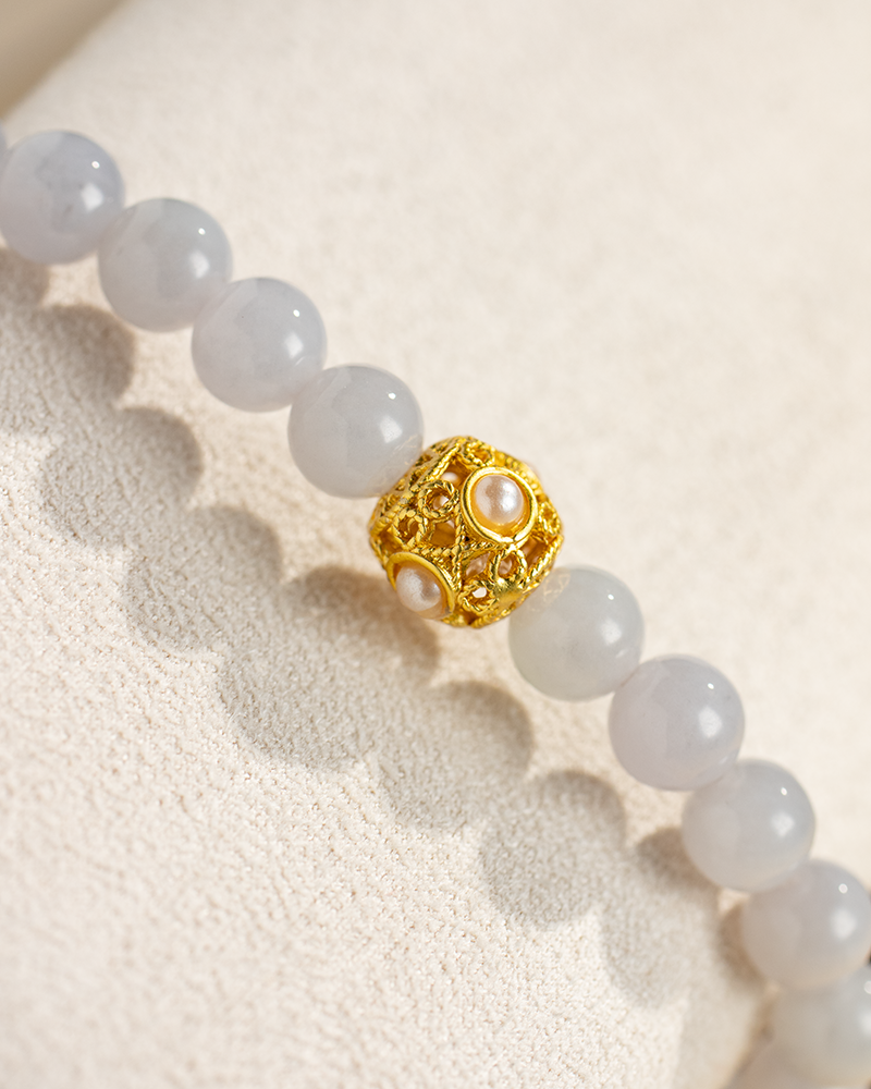 A close-up of a beaded bracelet with smooth, pale gray beads and a single ornate gold bead featuring small pearls, displayed on a textured light beige surface.