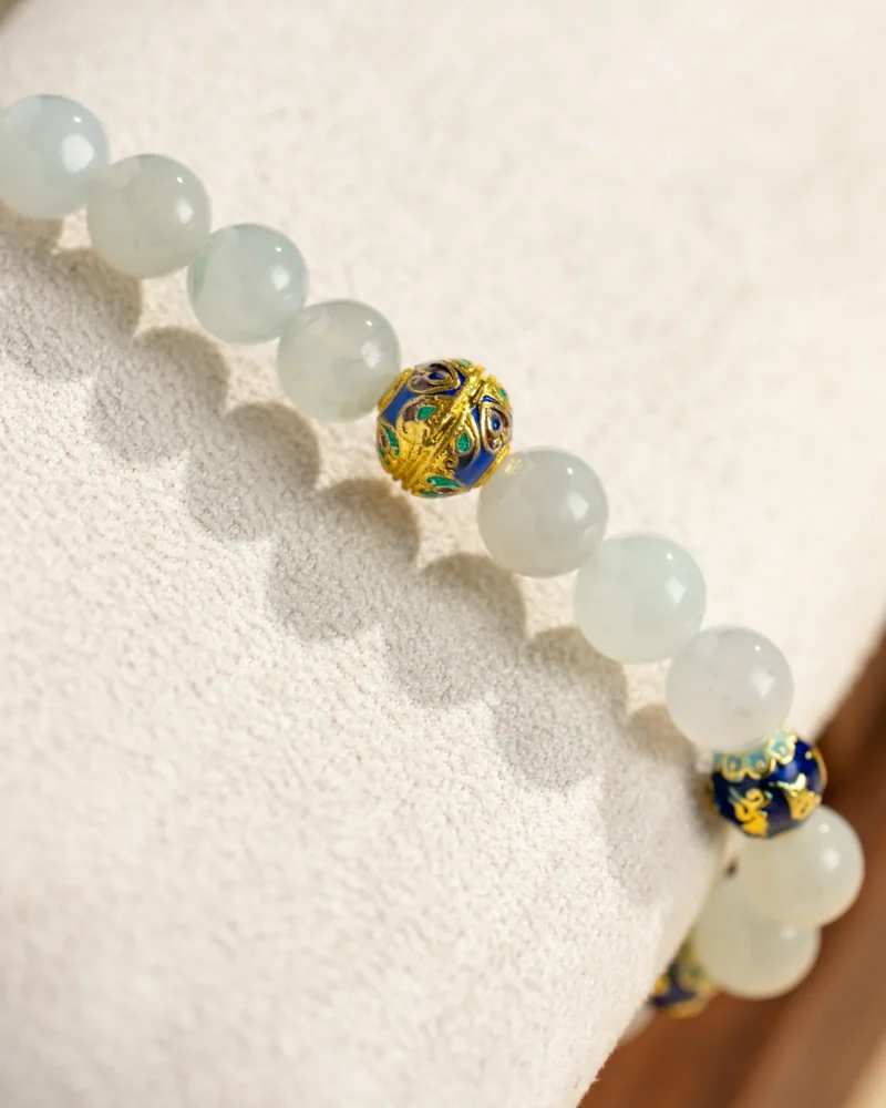 The Light Green Jadeite Bead Bracelet — Spherical Beads with Warm Accents is beautifully showcased on a soft beige textured surface, featuring pale green jadeite beads complemented by a single ornate blue and gold accent bead.