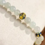 The Light Green Jadeite Bead Bracelet features pale green spherical beads with blue and gold warm accents, displayed on a soft, textured cream-colored surface.