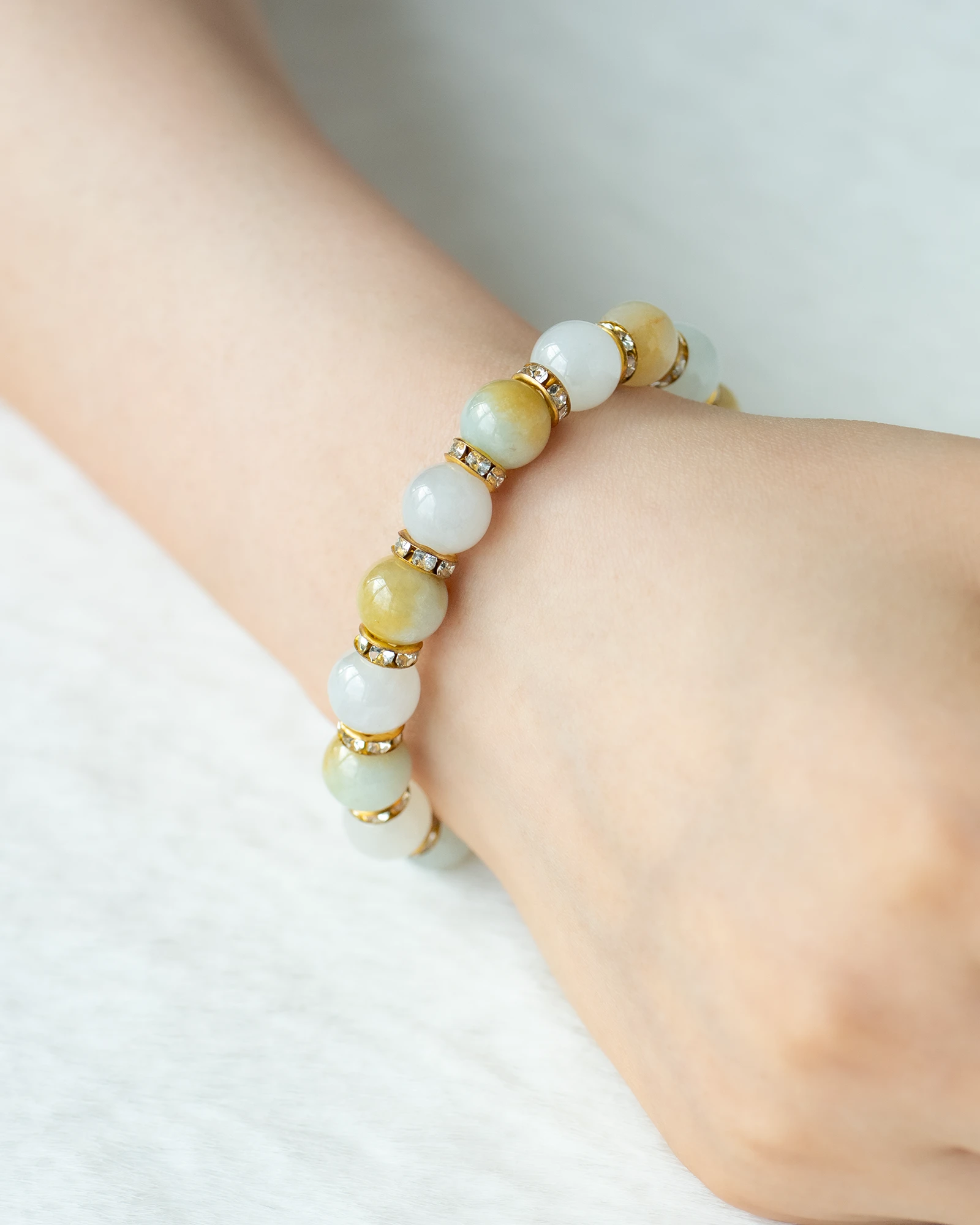 A close-up of a wrist adorned with the Greenish White and Brownish Yellow Jadeite Bracelet—featuring spherical beads accented by faceted details and separated by delicate gold spacers—highlights the refined elegance of jadeite jewelry against a soft, light background.