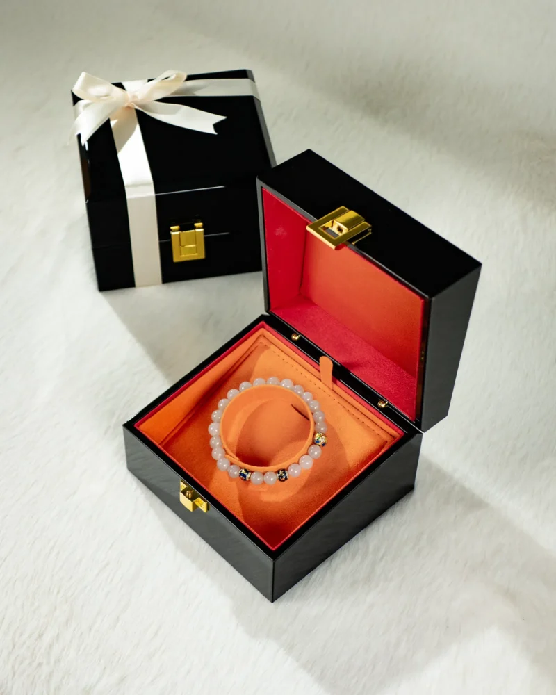 A black jewelry box with a gold latch, lined with orange fabric, holds the Light Green Jadeite Bead Bracelet—featuring spherical beads with warm accents. Another black box tied with a white ribbon rests in the background on a textured white surface.