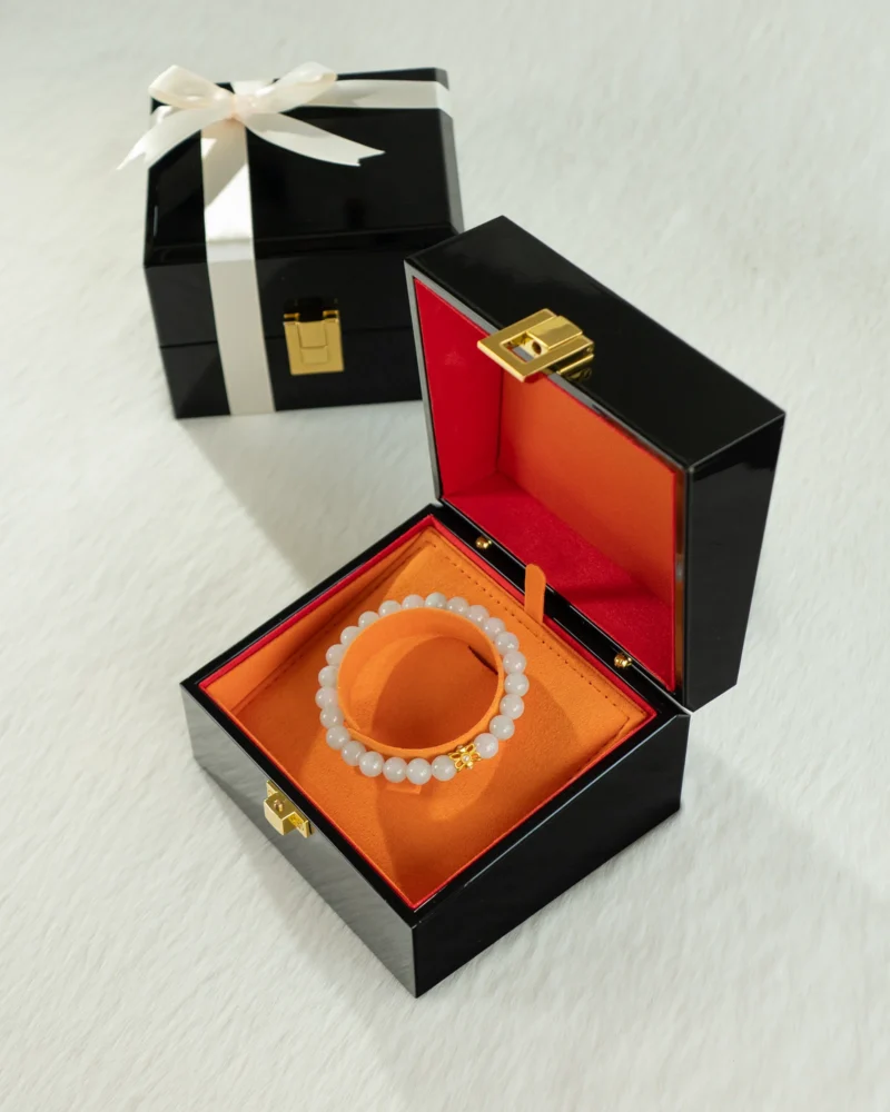 A Light Green Jadeite Bead Bracelet featuring spherical beads and a gold-tone detail is displayed in an open black and orange jewelry box. In the background, there is another closed black gift box tied with a white ribbon, both resting on a light surface.