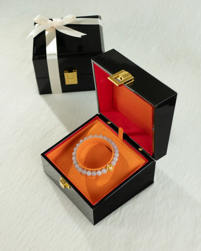 A Greenish-White Jadeite Bead Bracelet — Spherical Beads with Warm-Tone Accent rests elegantly in a black and orange jewelry box, accompanied by another closed black gift box tied with a white ribbon in the background, all beautifully arranged on a white textured surface.
