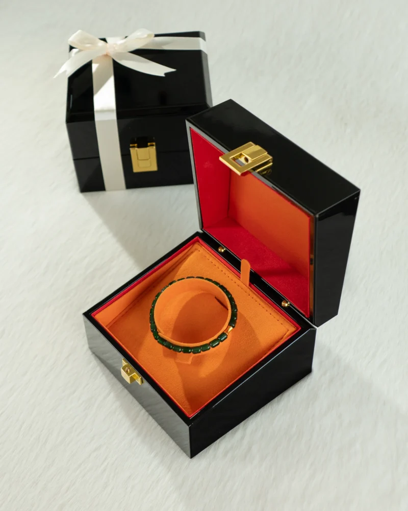 The Deep Green Feicui Bracelet – featuring drum-shaped beads with a warm-tone spacer – is showcased in an open black velvet box with an orange interior, while another closed gift box tied with a white ribbon sits in the background on a white surface.