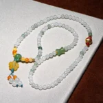 A beaded necklace with white, green, yellow, and orange beads, featuring a cluster of yellow beads and a decorative tassel, displayed on a white and brown textured surface.